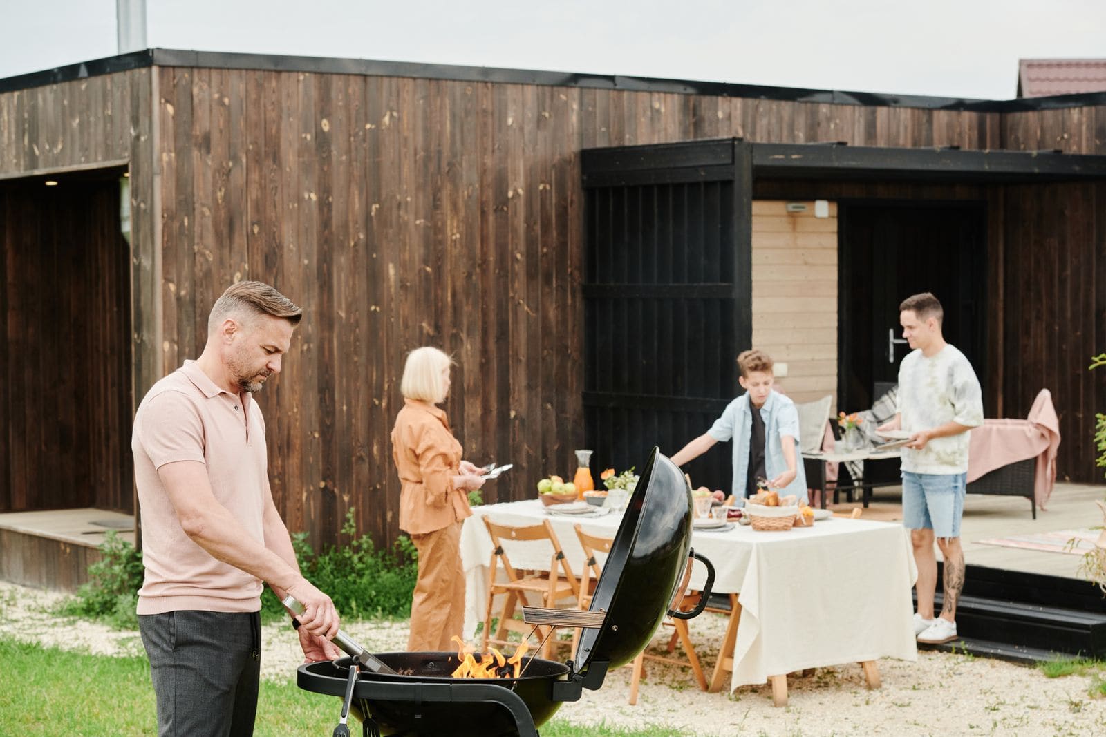 Man grilling on a backyard barbecue while family sets the table