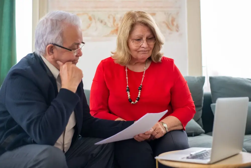 Older couple on a sofa reviewing paperwork beside a laptop